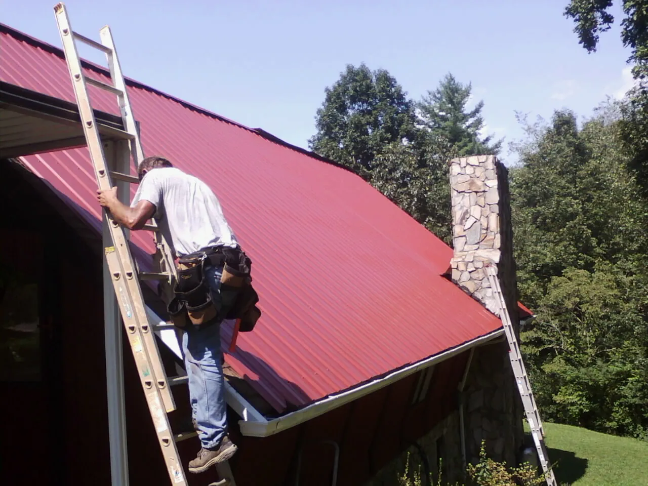 Roofer on a ladder beside a red metal roof and stone chimney — Soffit & Fascia Repair by Roofing Brothers in Centerburg